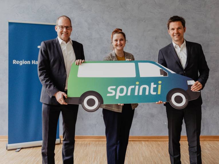 Drei Personen halten ein großes Pappfahrzeug mit der Aufschrift "sprinti" in den Händen